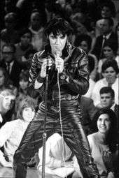 Presley, wearing a tight black leather jacket with Napoleonic standing collar, black leather wristbands, and black leather pants, holds a microphone with a long cord. His hair, which looks black as well, falls across his forehead. In front of him is an empty microphone stand. Behind, beginning below stage level and rising up, audience members watch him. A young woman with long black hair in the front row gazes up ecstatically.