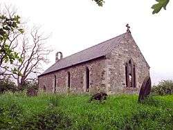 A photograph of a small stone church with open, glass-less windows. The church is surrounded by rough, uncultivated ground and the sky behind the church is bleak.