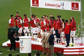 A coloured photograph of the Arsenal squad standing on a podium, celebrating their fourth Emirates Cup win.