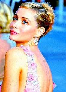A young blonde woman with her hair tied up at the back, wearing diamond earrings and a pink flowery dress, photographed from behind and to the left and looking sideways from the viewer