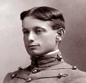 Head of a young man with neatly combed and parted hair wearing a military cadet's jacket, with cords running horizontally across the chest and a high, stiff collar.