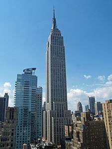 Aerial view of a 100-story building with several setbacks; the building tapers into a large circular spire near its 90th floor and is topped by a large antenna.