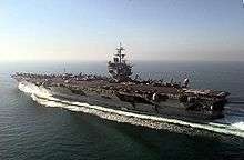 Aircraft carrier steaming away from camera in open sea. On deck is a large contingent of aircraft