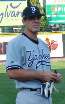 A man in a grey uniform and a navy blue hat holds a pair of white batting gloves.