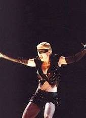 Madonna wearing black shorts and looking to her right. Her arms are open and a headset microphone to her mouth.