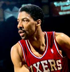 A basketball player, wearing a red jersey with the word "SIXERS" on the front, is looking to his right side.