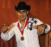 A dark man with black hat, white shirt, raising both hands, with a medal with red ribbon around his neck and legend BMI in the front.