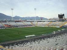 A semi-panoramic view of the stadium. The seats are white and black, and shows a the club badge in the seats. The roof of the stand is supported by a cantilever structure, in a "buried" construction.