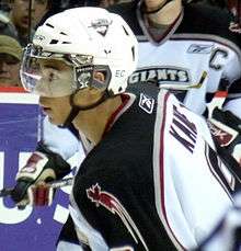 A black, teenage hockey player as seen from the side, cut off below the shoulders. He is looking towards the left intently during a game. He wears a white, visored helmet and a white, black and red jersey with "Kane" written on his upper back and a maple leaf on his upper arm.