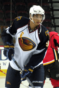 An African-Canadian hockey player. He wears a white jersey with dark blue shoulders with a stylized brown thrasher holding a hockey stick for a logo. He is standing with both hands on his stick with it on the ice.