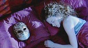 A woman sleeps on her bed. A mask lies on the pillow besides her.