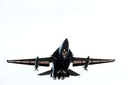 Low, head-on view of F-111 jet in flight, wings at minimum sweep with drop tanks underneath