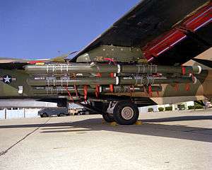 Closeup view of cylindrical bombs and ordnance carried under a mostly green aircraft wing