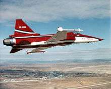 Starboard view of red and white single-engine jet fighter aircraft banking left. At the wingtips are missiles.
