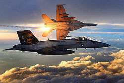 Two jet aircraft flying over clouds during dawn/dusk. The one in the foreground is perpendicular to the camera; the second further away is banking left while releasing orange flares