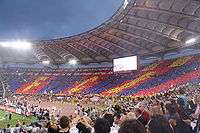 A full stadium, with fans holding up yellow, blue and red cards to make a giant mosaic.