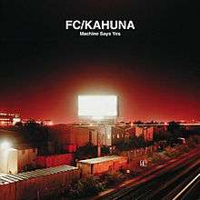 A nighttime photo showing distant buildings, bushes, railroad cars, and railroad tracks all under a billboard that is white and very illuminated, radiating out red light. Above the billboard is a line of uppercase text that reads "FC/KAHUNA", and underneath that is a smaller line of text that reads "Machine Says Yes".