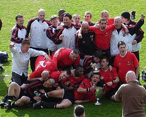 F.C. United players gather around the North West Counties League Division Two Trophy while fans take pictures.