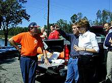 A group of eight middle-aged people surround the open back of a pickup truck that is carrying orange traffic cones and white food containers. The scene is next to a body of water and some trees and a telephone pole, on a blue sky sunny day. Three people are most prominent. One man is dressed in blue pants, and orange T-shirt from a hazardous materials team, and a blue and orange baseball cap. Another man is pointing at an unseen object, and is wearing blue jeans and a dark blue shirt, and has an air of authority about him. A woman is standing next to that man, listening. She is wearing black pants, a white shirt, and has brown hair cut above and down behind the ear and is wearing a thin black wristwatch.