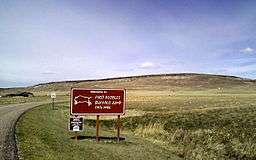A sign saying "Welcome to First Peoples Buffalo Jump State Park" in script type with a drawing of a buffalo next to it in white on a brown background. Behind it are some buildings and a large rise in the earth.