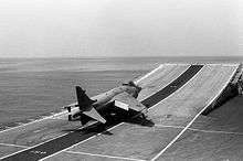 Black-and-white photograph of jet aircraft making a takeoff run at sea, approaching a ski-jump, which is a curved surface assisting aircraft in taking off