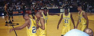 five Black males in gold athletic uniforms in the foreground on the sidelines of an athletic court while a few opposing athletes in green wait in the middle of the court.