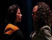 Two women are staring at each other while standing in a dark room. The woman on the right has ridges on her forehead and curly hair, while the one on the right has straighter hair and a uniform with a yellow strip.