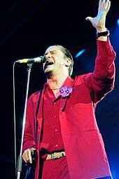 A man in a red suit standing in front of a microphone stand, with one arm raised