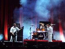 Three musicians dressed in white perform onstage—a drummer, a singer and a bass player