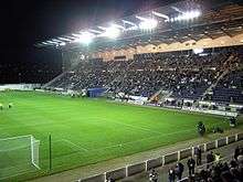 An evening game at a football stadium. The main stand on the right is filled with spectators and the empty pitch is being prepared for playing on the left