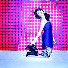 A young woman wearing a blue shirt kneels and poses during a photo-shoot, wearing zebra-printed pants, and high-top boots. The floor is metallic, and the background is filled with several small pink circles.