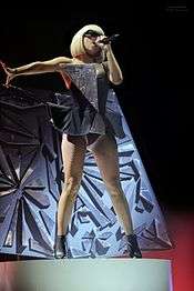 A blond woman standing on a stage. She wears a black tutu shaped dress with a shiny triangle attached on her right bosom. Her hair is in bob and she wears sunglasses while singing to a microphone held in her left hand. Behind her, a set piece encrusted with broken glasses is visible.