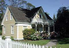 A modest two-story house sits behind a white wooden fence on a lot with a manicured lawn, blooming bushes, a sidewalk, and trees. The outside of the house, which is painted tan with white trim, looks new, as do the sidewalk and fence.