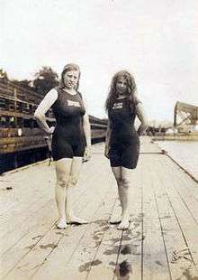 Two female swimmers stand on a wooden pool deck wearing bathing suits that have short sleeves and while full bodied, look like shorts go down to just above the knee