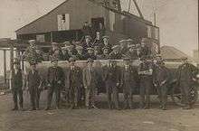 In a black and white photograph,  group of 26 young to middle-aged men wearing suits and flat caps stand in two rows. Behind them is a large, industrial shed, and in the distance the roof of a house can be seen.