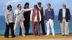 Seven men in suits standing near the ocean posing for a photo.