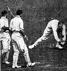 A cricket fielder picks up a ball, watched by two other players
