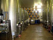 Colour photo showing stainless steel fermentation tanks in the UK.  The tanks are located on both sides of an aisle at the bottom of which oak barrels for aging, stacked on a bracket against the wall can be seen.  The floor is painted and pitched to a drainage channel in the middle of the winery.