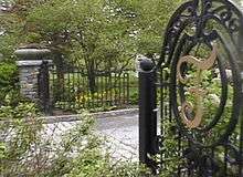 An open gate revealing the road to enter a cemetery, surrounded by grass, flowers and trees.