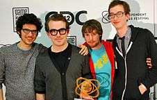 Four males from the development team stand with an atom-like trophy with a GDC backdrop. From left to right, Vreeland has long, messy hair, rectangular glasses, a TV static-like sweater, and is unshaven. His arm is around Fish, whose brown hair is combed back. He wears thick black glasses frames and a black shirt underneath a gray cardigan. McCartin has brownish-red hair and a goatee, and wears a Fez logo T-shirt underneath a red zippered hoodie. Bédard is a head taller than the bunch, and has short, brown hair, rectangular glasses, and is unshaven. He wears a black and white checkered dress shirt, a black tie, and a zippered, black hoodie.