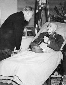 A man lies in a hospital bed, incongruously wearing an Army uniform instead of pajamas. His peaked cap is on the blanket and he holds a baton in his hand. A man in a dark suit and pinstripe trousers bends over to talk to him. In the background are flowers, and a flag.
