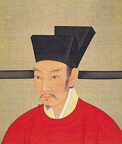A painting of a man with a black goatee looking to the left of the viewer while wearing a red shirt and a black hat, all in front of a grey background
