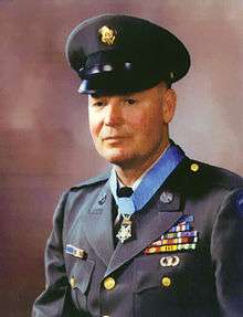 Head and shoulders of a white man wearing a peaked cap and a military jacket with yellow buttons and four rows of ribbon bars on the left breast. A star-shaped medal is hanging from a light blue ribbon around his neck.