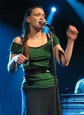 A woman wearing a green shirt, black belt and a dark gray skirt, standing behind a microphone on a stage.