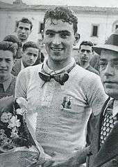 A man wearing a white jersey with goggles around is neck holding a bunch of flowers.