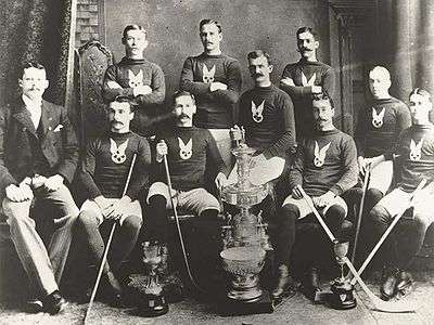 Two rows of ice hockey players, with the front row seated and most of the back row standing. Some of the players are holding hockey sticks, and various trophies are placed in front of them.