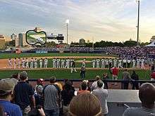 Baseball players in white uniforms with black caps line up along the third base line as they are introduced to the crowd at the ballpark