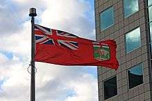 A red flag with the Union Jack in the top left corner and a coat of arms depicting a bison in the lower right corner flies beside a building
