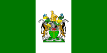A flag bearing three equally-wide vertical stripes, green, white, and green, with a coat of arms prominently emblazoned on the centre of the middle stripe.