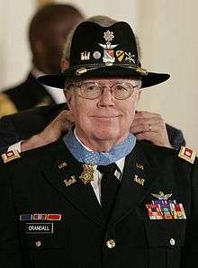 A color picture if Bruce Campbell in his dress military uniform and cavalry hat. He is smiling and President Bush can be seen putting the Medal of Honor around his neck.